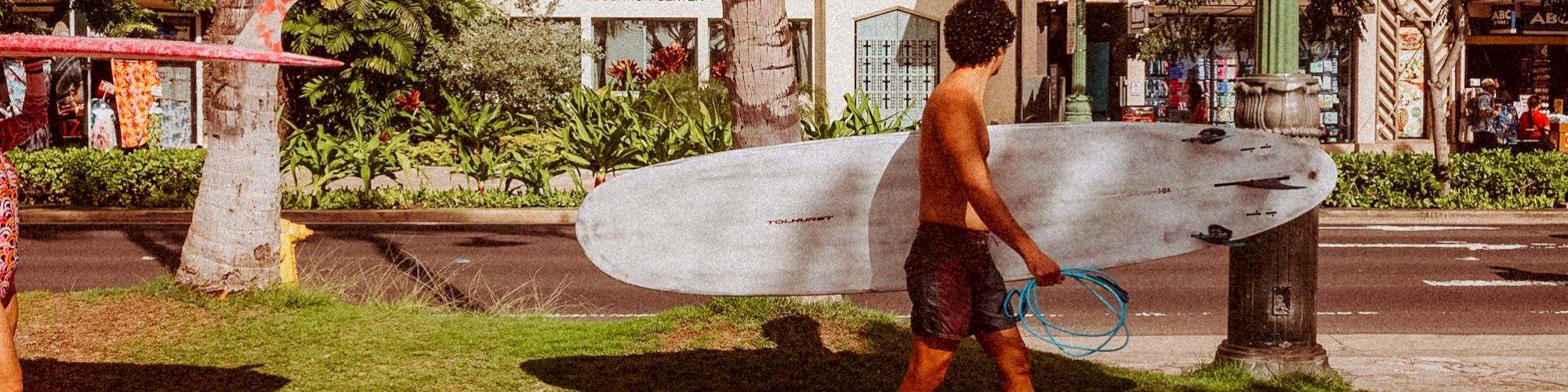 Two individuals are walking on a sidewalk, each carrying a surfboard under their arm, with palm trees and buildings in the background.