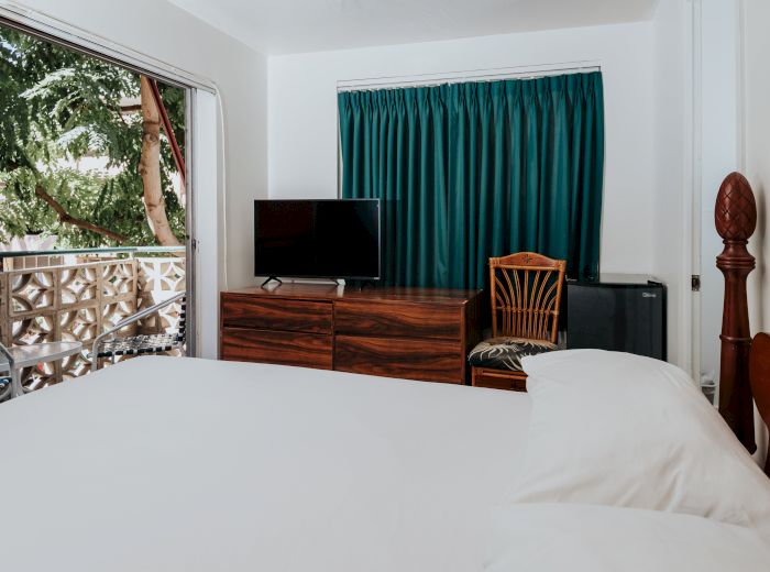 A cozy bedroom with a large bed, TV on a wooden dresser, a chair, green curtains, and a balcony with an outdoor view.