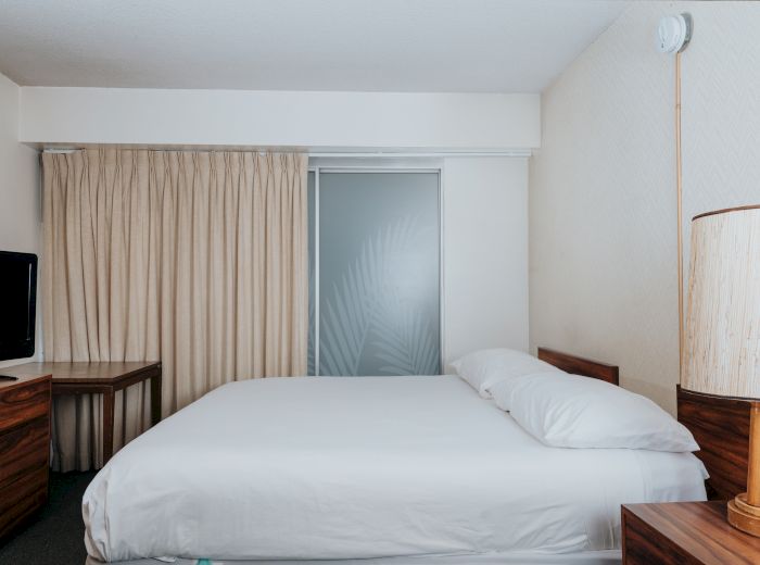 The image shows a minimalist hotel room with a bed, TV, desk, and lamp, neutral tones, and a frosted glass door with a leaf design ending the sentence.