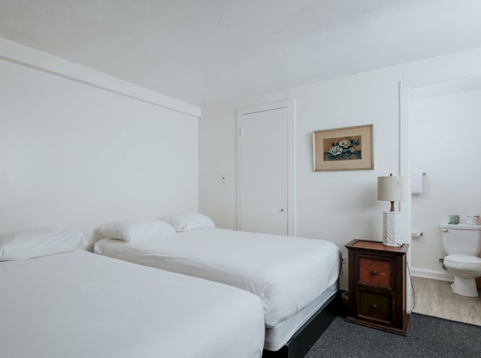 The image shows a clean and simple hotel room with two single beds, a bedside table with a lamp, a painting on the wall, and an adjacent bathroom.