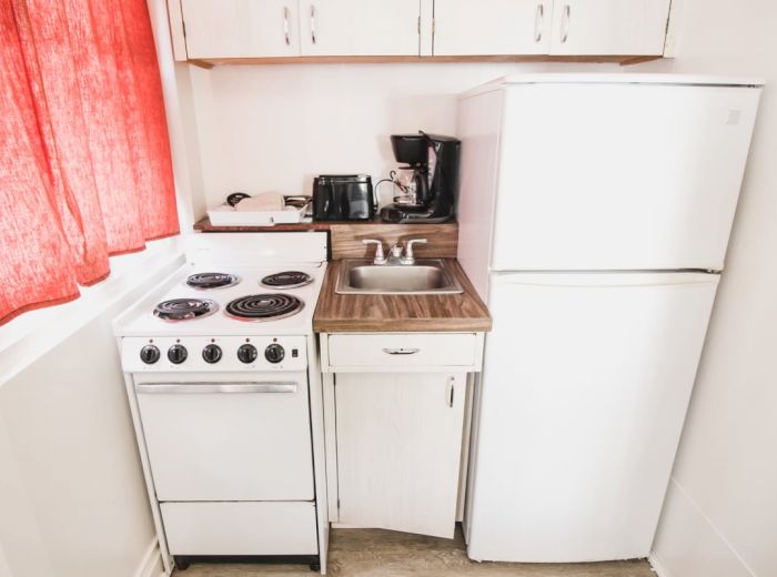 A compact kitchen with a fridge, stove, sink, toaster, coffee maker, white cabinets, and red curtains on a window.