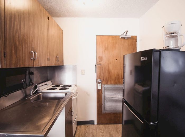 A small kitchen with wooden cabinets, a black refrigerator, a stove, a microwave, and a coffee maker on top of the fridge near a wooden door.