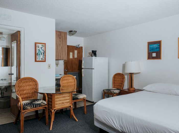 A hotel room with a bed, refrigerator, table with two chairs, a bathroom sink, two framed pictures, and a lamp on a side table.