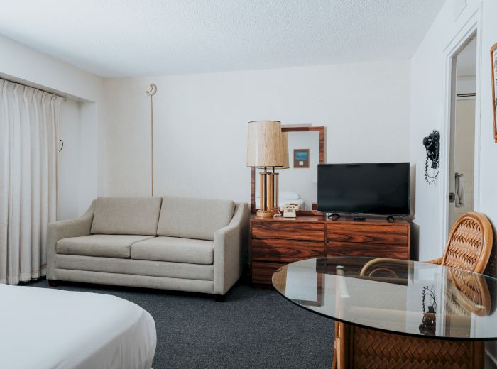 A hotel room features a sofa, a TV on a wooden dresser, a glass table with wicker chairs, and a bed partially visible on the left end of the sentence.