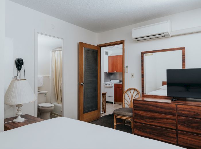A hotel room with a queen-sized bed, a bathroom, a TV, a wooden dresser, a mirror, an air conditioner, and a kitchenette in the background.