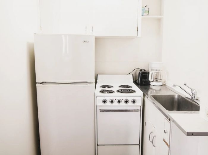 A small kitchen with a refrigerator, stove, sink, coffee maker, and some shelves. The setup is compact and efficient, suitable for limited space.