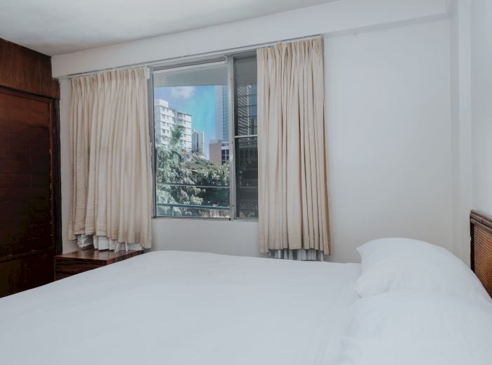 A cozy bedroom with a large bed covered in white linens, beige curtains on a window displaying a view of buildings outside, and wooden furniture.