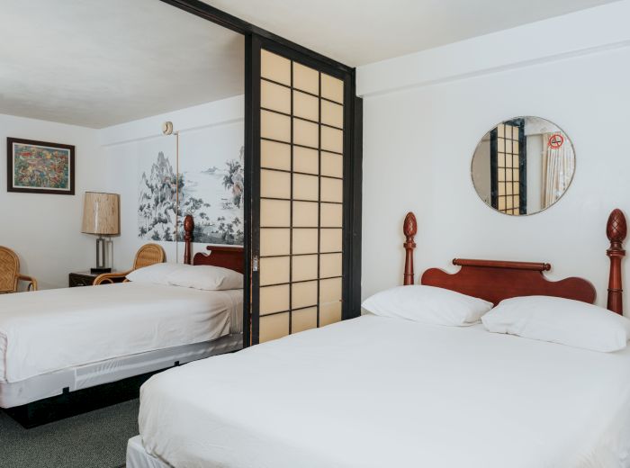 A hotel room with two double beds, a circular mirror, framed artwork, wicker chairs, a lamp, and a partition with Japanese-style sliding panels.