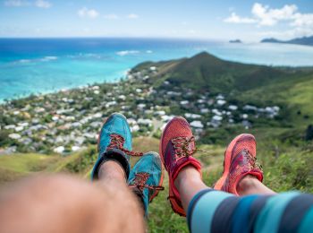 Two people with hiking shoes rest on a hill overlooking a coastal town with a view of the ocean and surrounding hills ending the sentence.