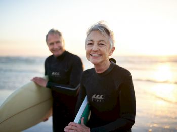 Two people in wetsuits, holding surfboards, standing on a beach with the ocean and sunset in the background, looking at the camera.