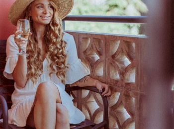 A woman with long curly hair and a wide-brimmed hat sits on a balcony, holding a glass, wearing a white dress.