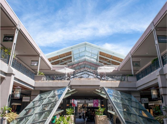 The image shows the entrance of a modern shopping mall with escalators, glass roofing, and various store signs under a blue sky with clouds.