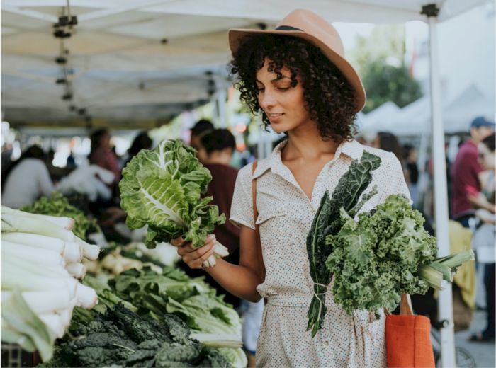 A woman is at a farmer's market, holding fresh leafy greens and wearing a hat. She is surrounded by various vegetables displayed on the stall tables.