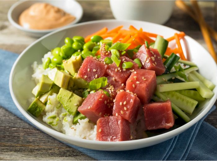 A bowl of poke with tuna cubes, avocado, edamame, cucumber, carrots, on a bed of rice, garnished with green onions and sesame seeds, and sauce on the side.