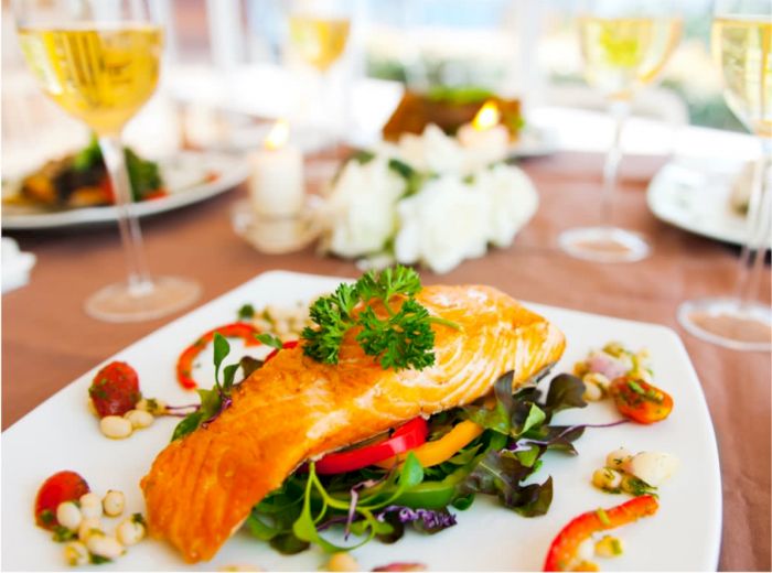 A plated dish featuring grilled salmon on a bed of mixed greens with garnishes, surrounded by glasses of white wine on a set dining table.