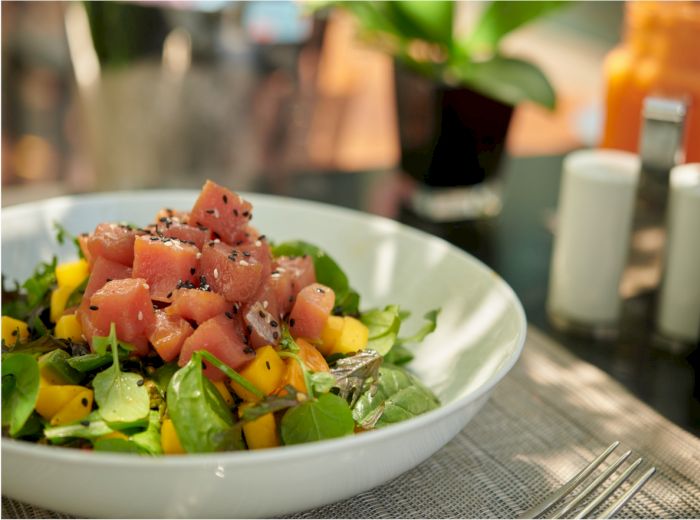 A salad features fresh greens, diced raw tuna, mango chunks, and sesame seeds, with salt and pepper shakers and a plant nearby, on a table.