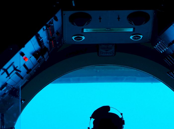 A person is inside a vessel, viewed from behind, looking out a large circular window into a blue environment, possibly underwater, wearing a headset.