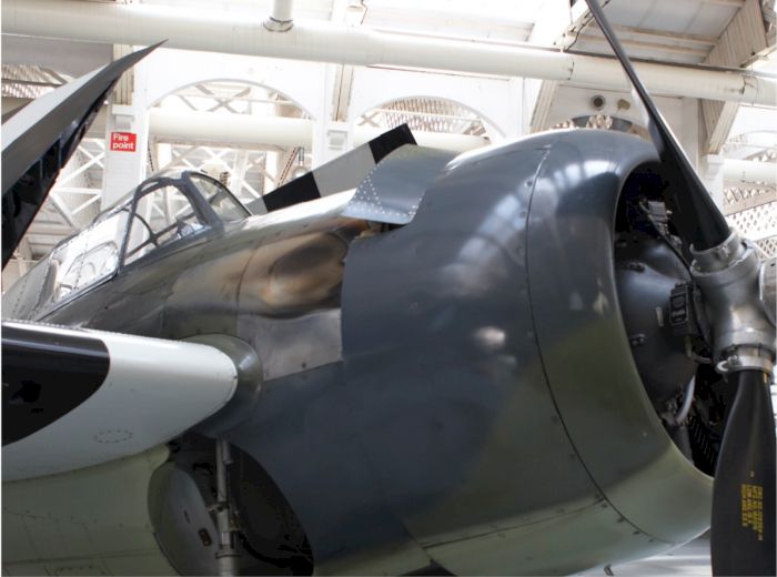 The image shows a close-up view of a vintage aircraft, focusing on its cockpit and propeller, inside a museum or hangar.