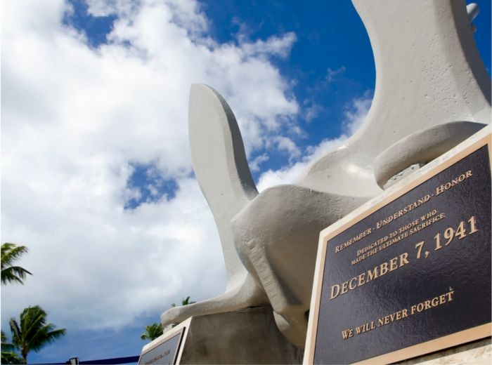 The image shows a memorial plaque dated December 7, 1941, with the inscription "WE WILL NEVER FORGET," against a background of a partly cloudy sky.