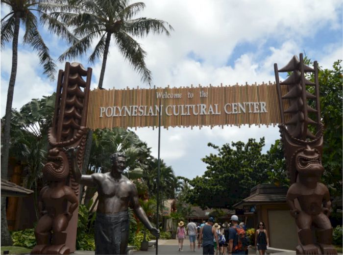 The image shows the entrance to the Polynesian Cultural Center, featuring a sign and statues, with palm trees and people in the background.