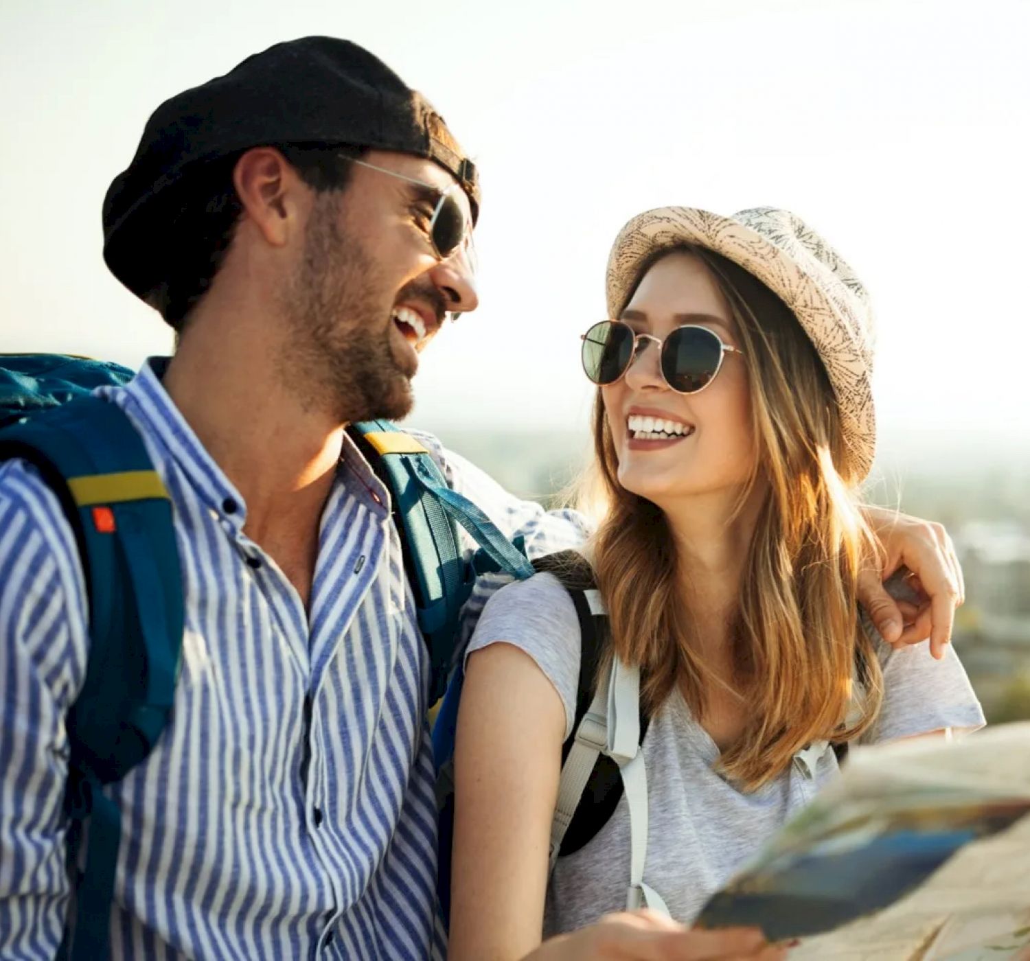 A smiling couple wearing sunglasses and backpacks looks at a map outdoors. They appear to be enjoying a sunny day of exploration.
