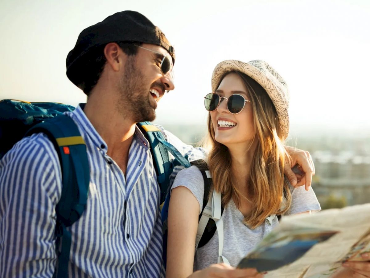 A smiling couple wearing hats and sunglasses, carrying backpacks, looking at a map while enjoying a sunny day outdoors.