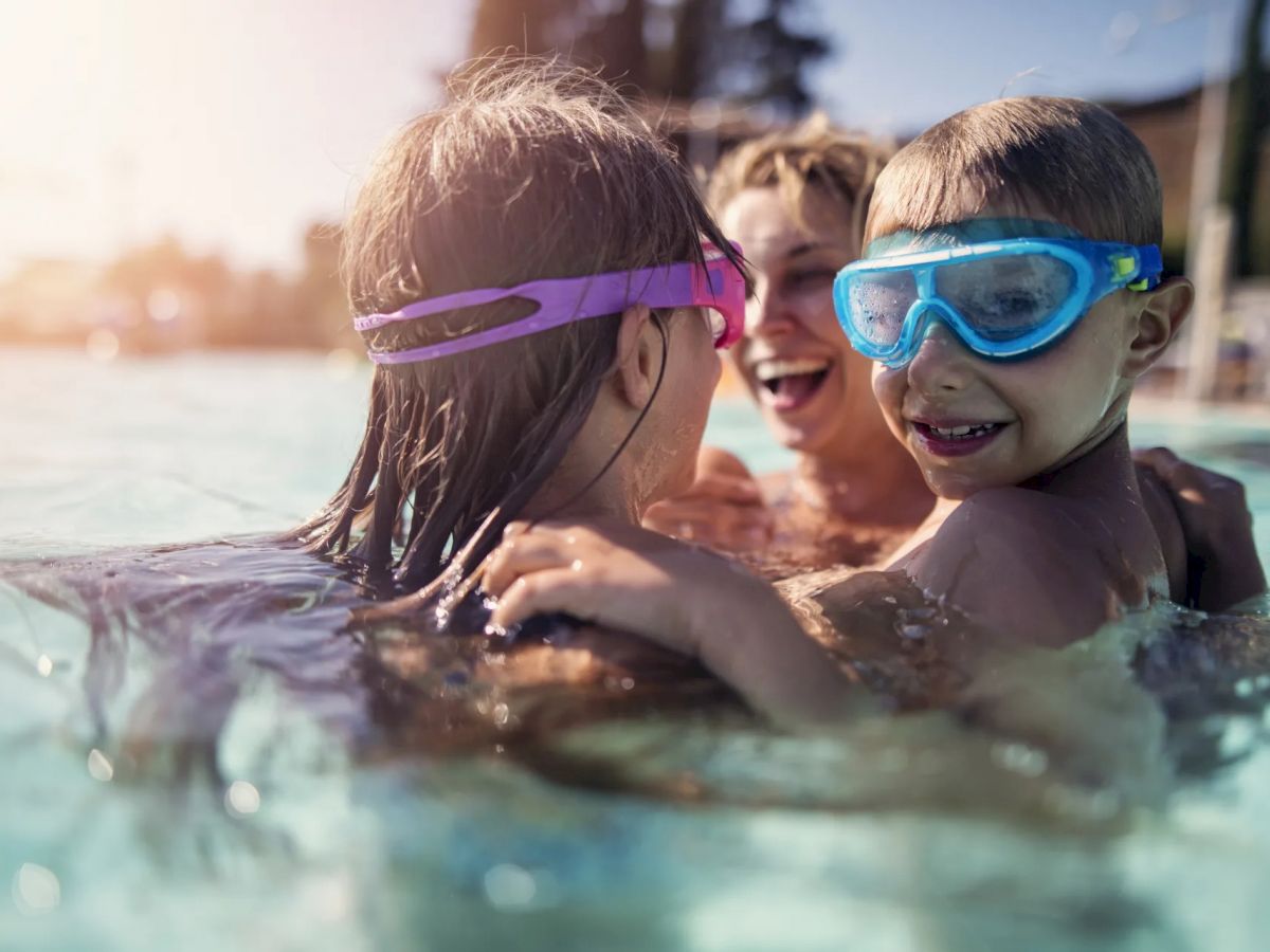 Three people, two children and an adult, are enjoying their time in a swimming pool, wearing goggles and smiling widely.