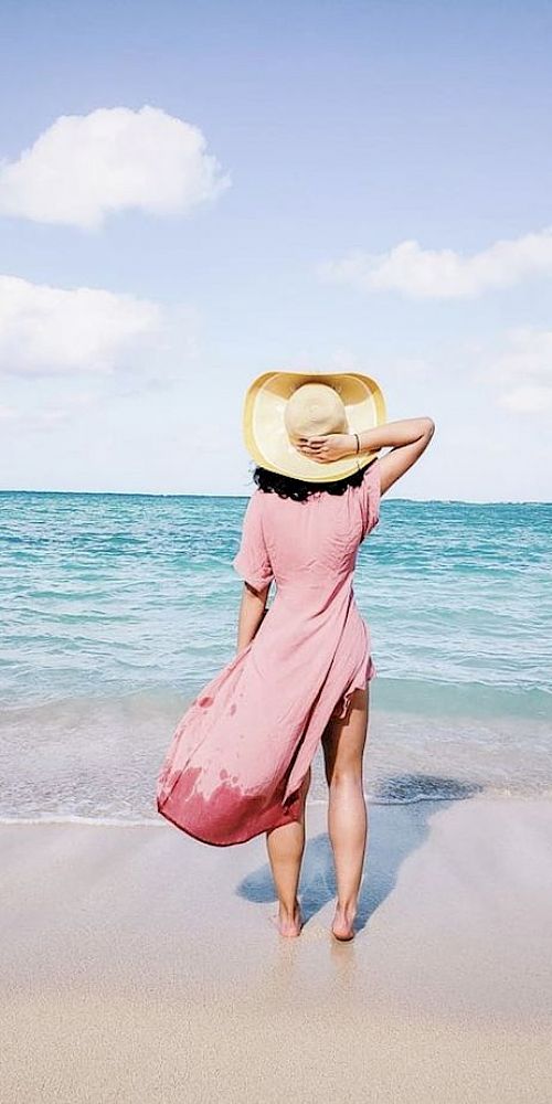 A person in a pink dress and yellow hat stands on a sandy beach facing the ocean with a blue sky and clouds in the background.