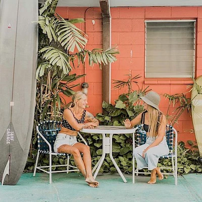 Two people sit at a table outside, surrounded by plants and surfboards against a coral-painted wall, with one window and two doors in view.