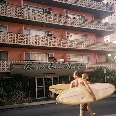 The image shows two people carrying surfboards walking past the Royal Grove Hotel, a multi-story building with balconies.