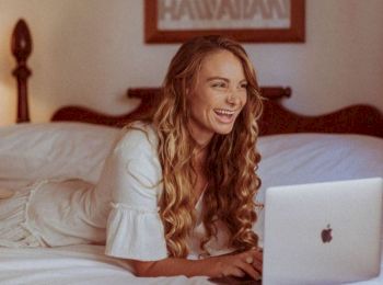 A woman with long hair is lying on a bed and smiling while using a laptop. There is a framed picture on the wall behind her.