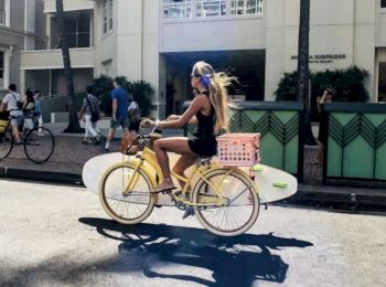 A person rides a yellow bike carrying a surfboard and a pink basket on the back, while people walk and bike in the background.