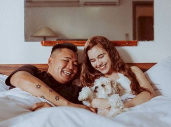 A couple is lying in bed, smiling and cuddling with a small dog.