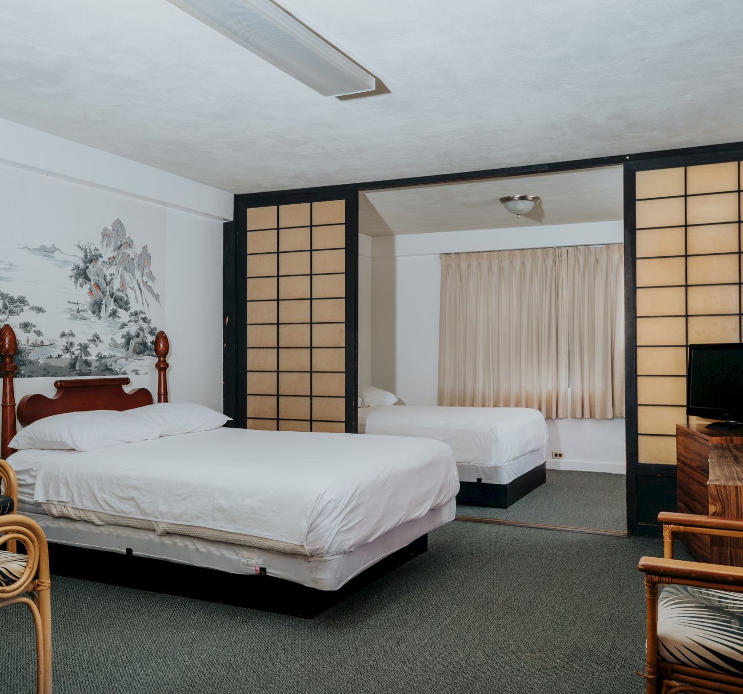 The image shows a hotel room with two beds, wooden furniture, chairs, a small TV, and a partition wall with a painting. The room appears cozy.