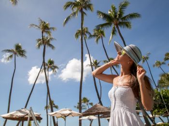 A woman in a white dress and sun hat enjoys a sunny day, surrounded by palm trees and blue skies, with beach umbrellas in the background.