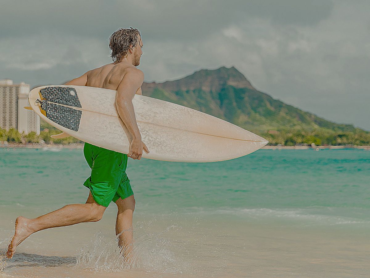 A person in green shorts runs on the beach with a surfboard, towards the ocean, with buildings and a mountain in the background.